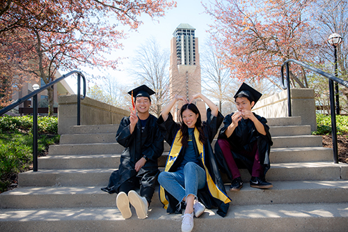 3 students in grad cap and gowns sit on steps outside in front of bell tower in spring