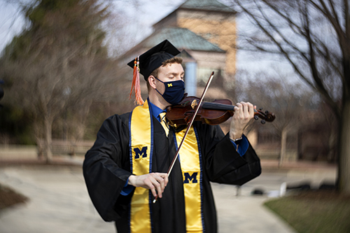 Matthew Lamb in grad cap, gown, sash, and mask plays the violin on North Campus